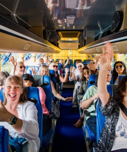 A front-view shot of a large multi-ethnic group of tourists celebrating on a coach bus, they are smiling and raising their arms with excitement, they are ready to begin their journey.